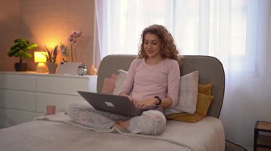 Genç bir kadın yatağının konforundan dizüstü bilgisayarıyla çalışıyor, pijama giyiyor.