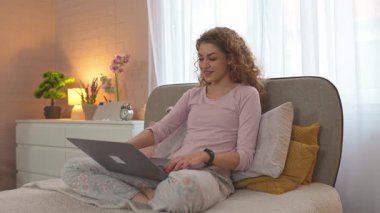 Pijamalı güzel bir kadın laptopuyla çalışıyor ve yatak odasındaki telefona cevap veriyor.