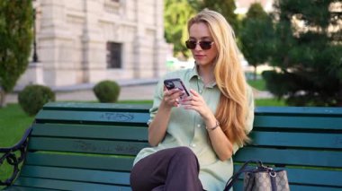 Yeşil bir bankta oturan gülümseyen genç bir kadın cep telefonuyla sosyal medyada geziniyor.