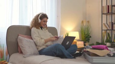 Genç bir kadın açık bir bavulun yanında yatağında otururken dizüstü bilgisayarından uçak bileti alıyor.