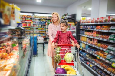 Anne ve kızı süpermarket koridorunda içi taze ürünlerle dolu bir alışveriş arabasını itiyorlar.