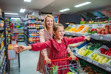 Genç bir kız alışveriş arabasında giderken annesi süpermarkette itiyor.
