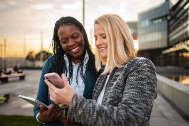 İki gülümseyen iş kadını akıllı telefonu kontrol ediyor ve şehir merkezinde bir tablet tutuyor.