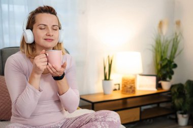 Kulaklık takan, gözleri kapalı, müzik dinlerken sıcak bir içki içen bir kadın.