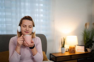 Kadın gözlerini kapatıyor, kulaklıkla müzik ve kahveden zevk alıyor.