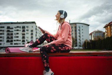 Woman sitting on bench with yoga mat, looking away while listening to music