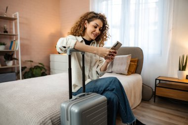 Bavullu gülümseyen kadın modern yatak odasında otel rezervasyonu için akıllı telefon kullanıyor.