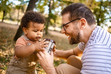 Bir baba ve küçük çocuğu canlı bir parkta neşeli bir anın tadını çıkarıyorlar. Güneş ağaçların arasından süzülür, yapraklarla çevrili renkli bir fincanın üzerinden geçerler..