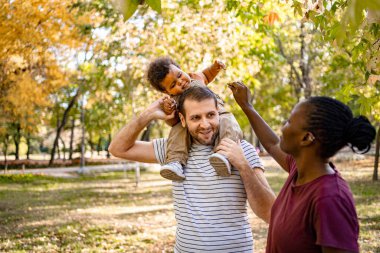 Sonbahar yapraklarıyla süslenmiş canlı bir parkta, bir baba çocuğunu neşeyle omuzlarında taşırken, bir anne onlarla neşeyle etkileşime geçer, doğada sevgi dolu aile anları yaratır..