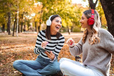 İki kız, canlı sonbahar yaprakları altında sıcak çimenlerde oturup kahkahalarını ve müziğini paylaşıyor. Neşeleri serin günü aydınlatıyor.