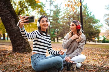 İki kız yere düşmüş yaprakların arasına oturmuş, gülümsüyor ve selfie çekiyor. Sonbahar renklerine bürünmüş ağaçlar neşeli sahnelerinin etrafını sarıyor..
