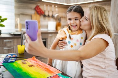 Bir anne, mutfaktaki sanat oturumunda mutlu bir şekilde selfie çekerken kızını öper..