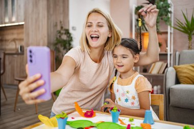 Neşeli bir anne-kız selfie için poz veriyor, masada canlı modelleme kil kreasyonlarıyla çevrili..