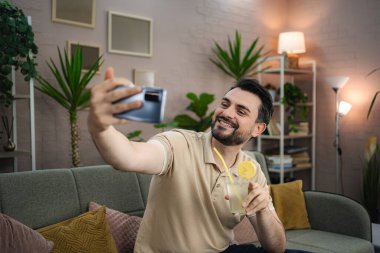 Oturma odasında limonata içerken akıllı telefonuyla selfie çeken mutlu genç adam.
