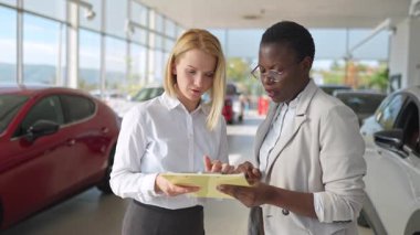 İki çok ırklı iş kadını modern bir araba galerisinde bir sözleşmeyi tartışıyorlar.
