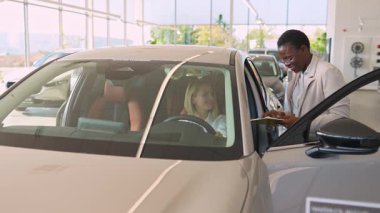 Bir danışman araba galerisinde belgeleri imzalarken mutlu bir kadın yeni bir arabada oturuyor.