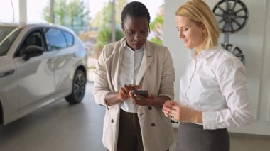Afro-Amerikalı bir satış temsilcisi ve beyaz bir bayan müşteri bir araba galerisinde akıllı telefon kullanıyorlar.