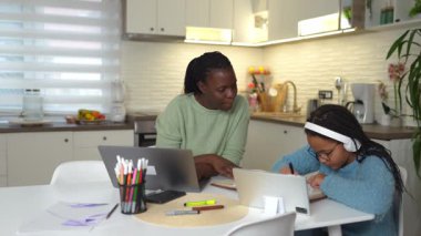 Anne, küçük oğlu mutfak masasında oynarken kızına ev ödevlerinde yardım ediyor.