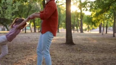 Neşeli anne, sıcak bir akşamda güneşli bir parkta gülen kızını elleriyle çeviriyor.