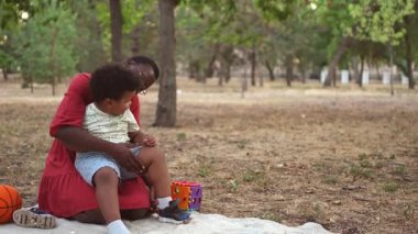 Siyah anne piknik battaniyesinde kucağında küçük oğluyla halka açık bir parkta oturuyor.