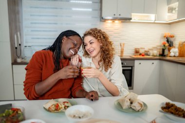 İki farklı kadın arkadaş yemek yiyor, kadeh tokuşturuyor ve modern bir mutfakta gülüyorlar.