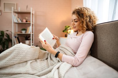 Genç bir kadın yatakta rahat rahat kitap okuyor.