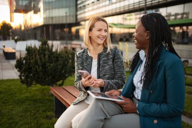 İki farklı iş kadını dışarıda bir bankta otururken gülüyor ve paylaşıyor.