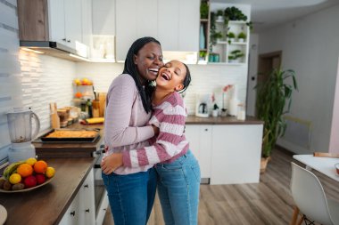 Anne ve kızı, modern bir mutfakta neşeli bir kucaklaşma ve kahkaha paylaşıyorlar.