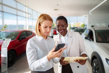 Two women discussing car features and prices on a smartphone in a showroom