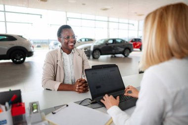Happy customer smiling while sitting at car dealership with saleswoman using laptop