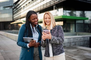 İki iş kadını bir akıllı telefonu paylaşıyor ve modern bir ofis binasının önünde gülümsüyor.
