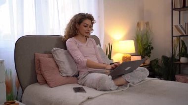 Genç bir kadın pijamalarıyla yatakta oturuyor, dizüstü bilgisayarında daktilo yazıyor ve uzaktan çalışıyor.