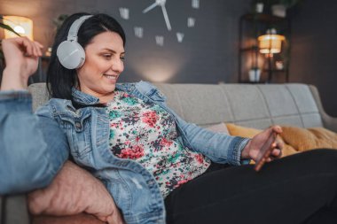 Kadın kulaklıkla müzik dinlerken telefonunu kullanıyor ve evdeki kanepede dinleniyor.