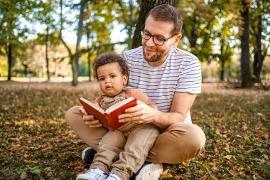 Canlı bir sonbahar parkında bir baba, renkli yapraklarla çevrili bir kitap okuyarak çocuğuyla sevgi dolu bir anı paylaşır. Ailenin sıcaklığı aşikar..