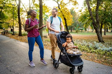 Bir çift, altın sonbahar yapraklarıyla çevrili canlı bir parkta neşeli bir yürüyüşün tadını çıkarıyor. Baba bebek arabasını iterken anne kahveyi yudumlar ve mutlu bir şekilde sohbet eder..