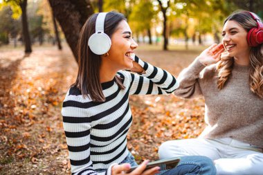 İki kız, neşeli bir sonbahar parkındaki renkli ağaçlarla çevrili, düşen yaprakların üzerinde otururken kahkaha ve müziği paylaşıyor..