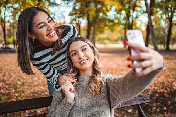Two girls smile brightly as they take a selfie in a park filled with vibrant autumn leaves, sharing laughter and friendship on a sunny day.