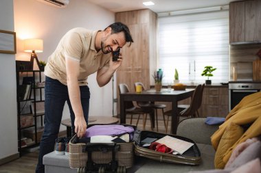 Gülümseyen adam telefonda konuşurken bavula giysilerini yerleştiriyor, yolculuğa hazırlanıyor.