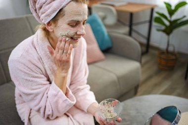 Bornoz ve havlu giymiş bir kadın yüzüne canlandırıcı kilden bir maske takıyor.