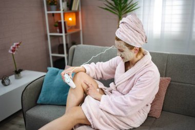 Kanepede oturan genç bir kadın, bornoz ve yüz maskesi takıyor, evde saç aldırmak için modern bir lazer epilatör kullanıyor.