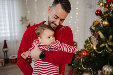 Baba kucağında bebek, Noel ağacına bakıyor, tatilini evde kutluyor.
