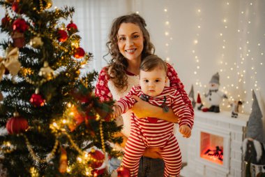 Süslü Noel ağacının yanında pijamalı bebeği tutan mutlu anne.