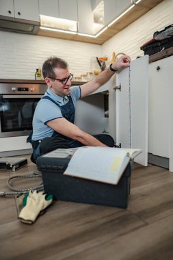 Mutfak dolabını ölçen tamirci yanında açık bir alet çantasıyla yerde yatıyor.