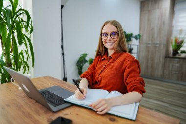 Rahat bir çalışma alanında gülümseyen kadın laptopuyla not alıyor..