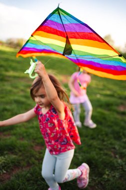 İki genç kız neşeyle yeşil bir tarlada koşuyor, biri canlı bir uçurtma taşırken kahkahaları havayı dolduruyor. Güneş parıldıyor, dışarıda geçirdikleri keyifli deneyimleri arttırıyor..