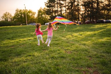 İki genç kız ellerinde canlı çizgili bir uçurtmayla yeşil bir alanda neşeyle koşuyorlar. Sıcak güneş ışığı manzarayı altın renginde yıkar ve neşeli bir atmosfer yaratır..