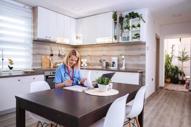 Doktor mavi önlük giyip evden çalışıyor, ahşap masada oturuyor ve tıbbi kayıtları kontrol ediyor.