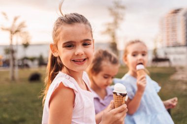 Üç anaokulu kızı güneşli bir yaz gününde parkta dondurma külahı yiyor.