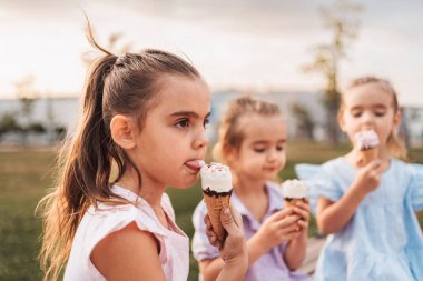 Parkta güneşli bir günde üç küçük kız birlikte dondurma yiyorlar.