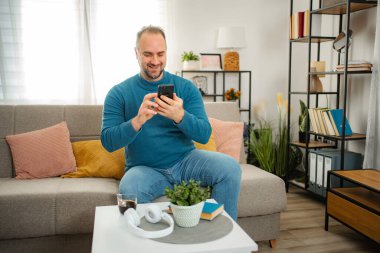 Adam kanepede otururken gülümsüyor, oturma odasında akıllı telefonuyla yazı yazıyor.